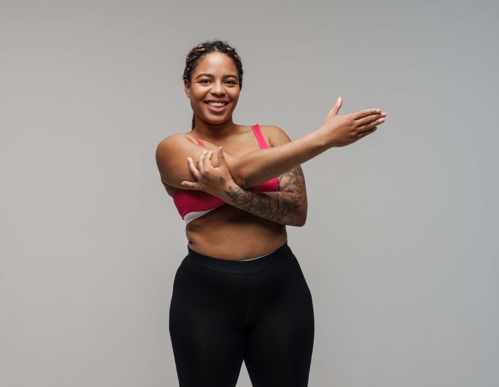 Smiling young african bodypositive fitness woman over gray wall background stretching exercise